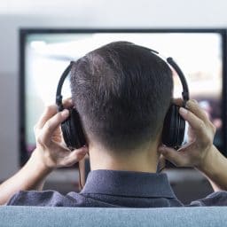 Back side of a man wearing black headphones in front of blurry out-of-focus television and home entertainment system background