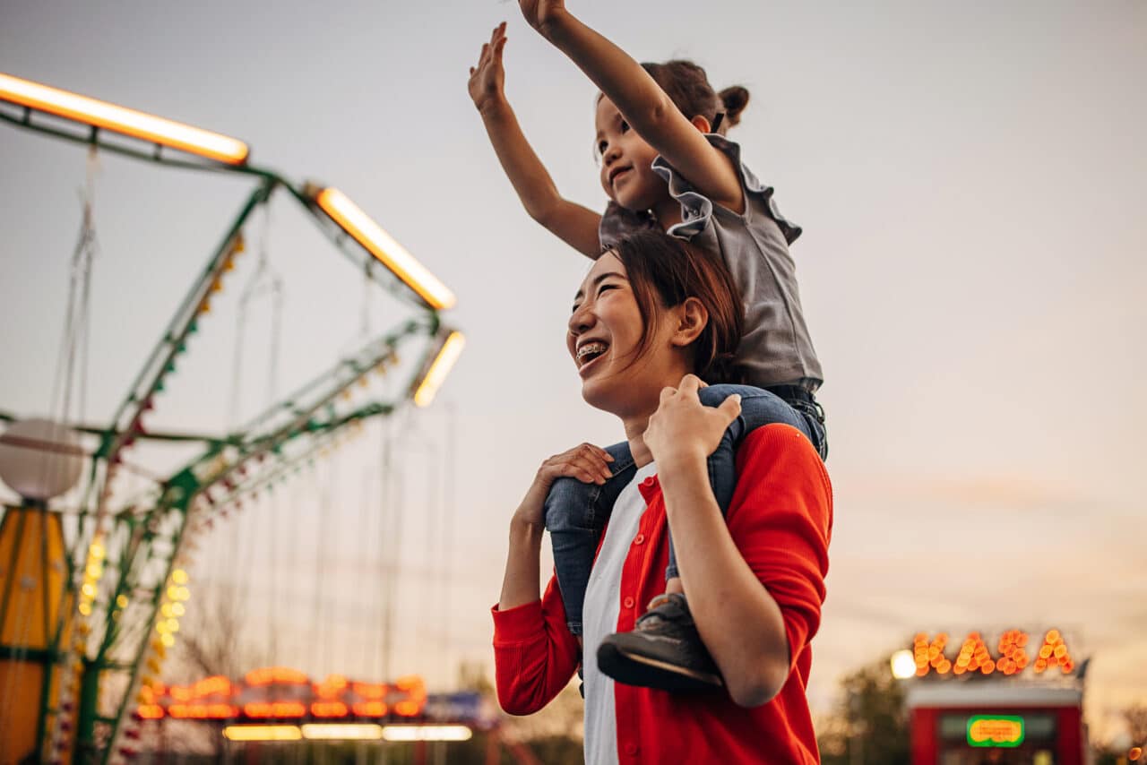 A mother carries her cut daughter on shoulders at amusement park.