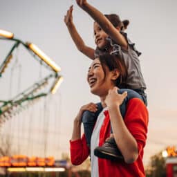 A mother carries her cut daughter on shoulders at amusement park.